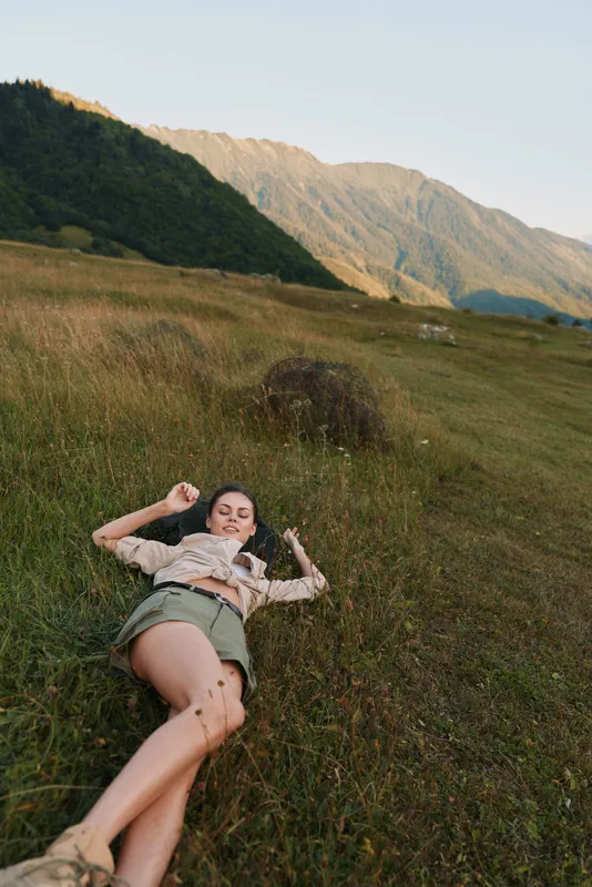 Woman lifestyle outdoors, lying in grass on a sunlit mountainside, relaxed pose amid meadow and natural landscape, gentle breeze, warm light, cheerful mood
