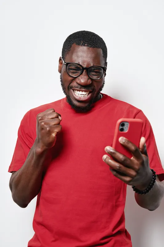 African American man with dark skin wearing glasses and red shirt celebrating success while looking at smartphone with clenched fist and joyful expression on white background
