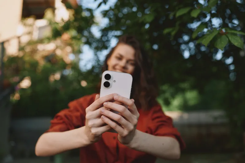 Smiling woman outdoors taking a selfie with a smartphone, casual red outfit, natural daylight, lifestyle scene emphasizing wellness, connection, and cheer