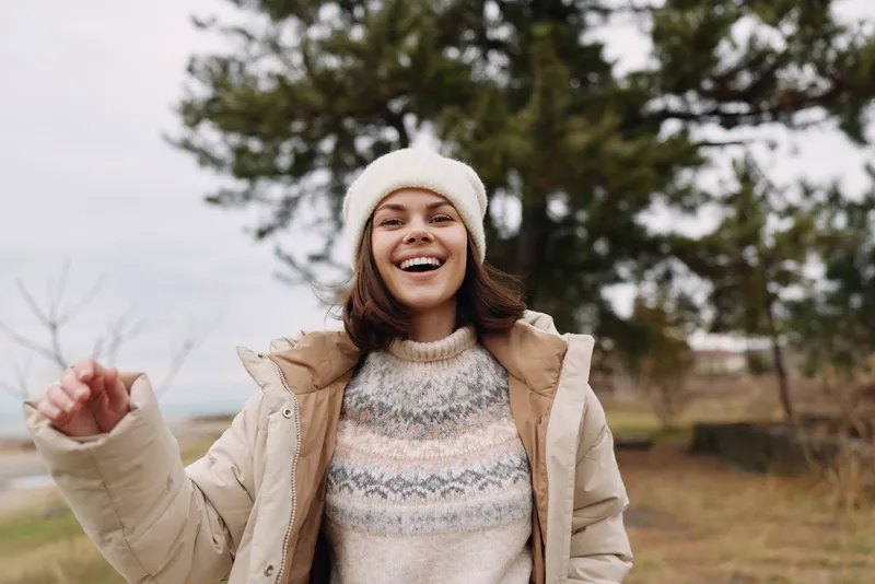 Happy woman in winter outdoor scene wearing a warm coat, knit sweater and beanie, enjoying fresh air and sunshine, posing with arms raised in a carefree moment.