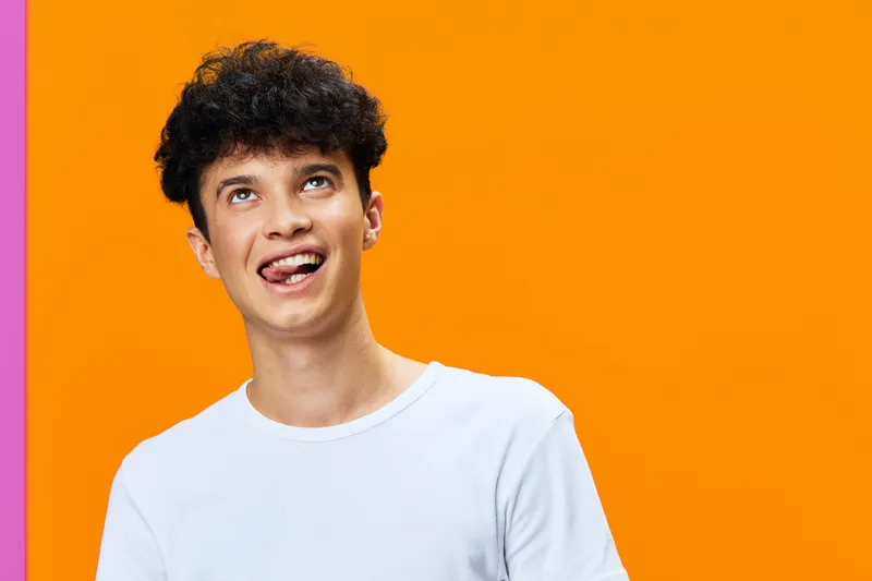 Young man with curly hair in white shirt making playful face against bright orange background expressing fun and creativity in casual style.