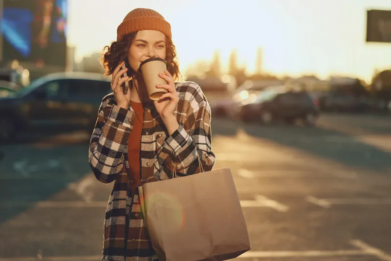 Woman drinking coffee and talking on phone while shopping at sunset, wearing beanie and holding paper bag, candid smiling portrait with golden hour glow, authenticity and mindful living.