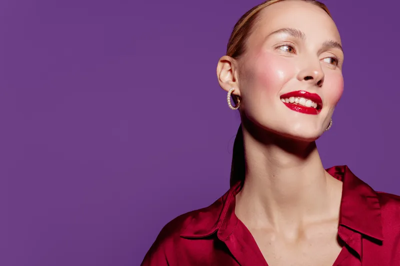 Smiling woman with red lipstick and earrings looks away on purple background. Portrait of confident female model in red blouse with natural skin and bright makeup. Beauty and fashion concept.