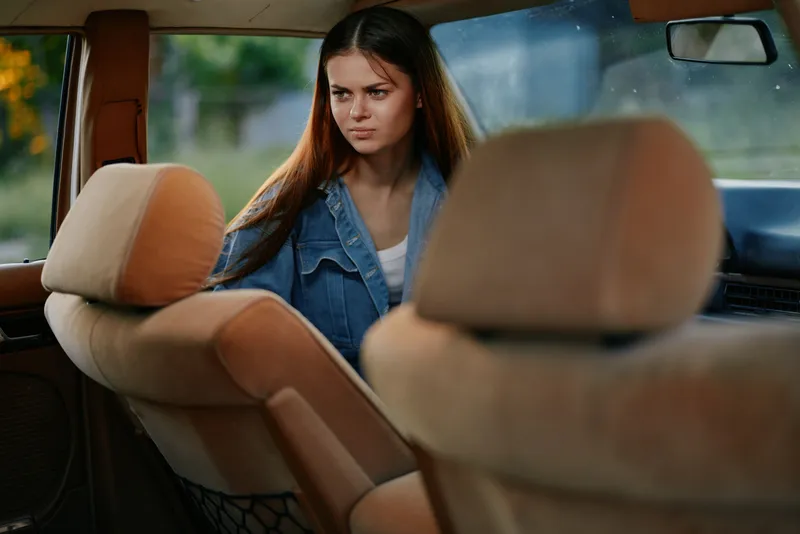 Young woman with long hair in denim jacket, showing contemplative mood inside an old car, highlighting vintage style and emotional depth.