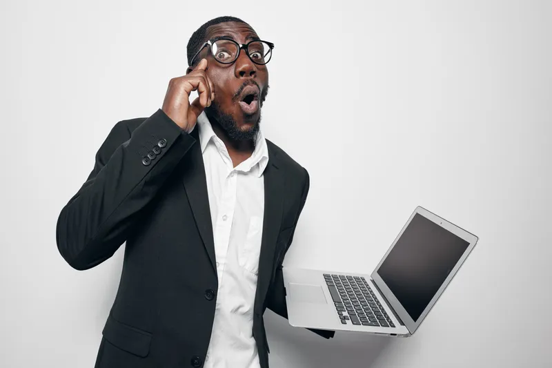 African American man with dark skin and glasses holding laptop with surprised expression in studio. Business casual dressed male showing shock or amazement with technology device on white background.