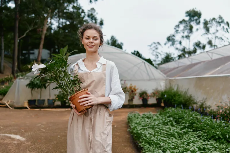 Young woman in an apron holding a potted plant, smiling in a greenhouse, surrounded by greenery and natural light for botanical themes