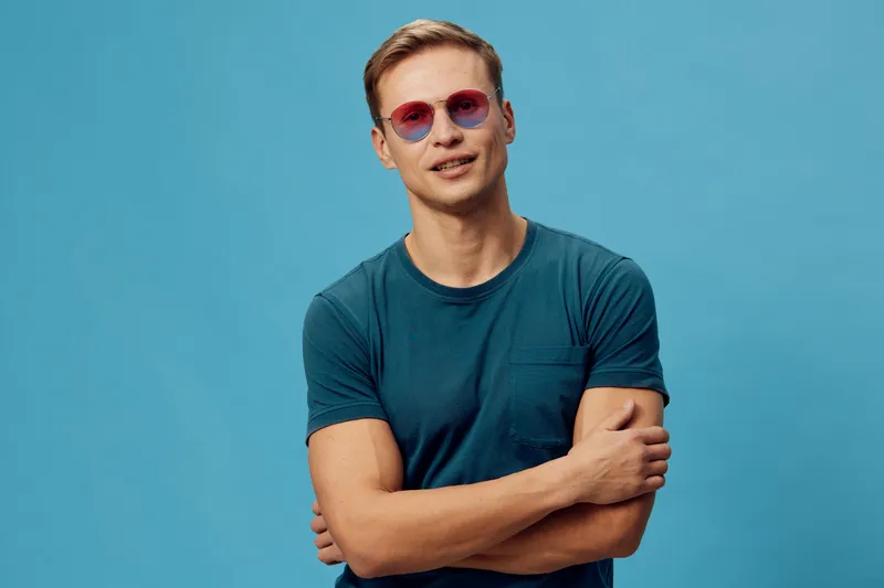 Casual young man with short hair wearing tinted pink sunglasses and teal t-shirt poses with arms crossed against plain blue background, relaxed expression, studio portrait. People lifestyle concept