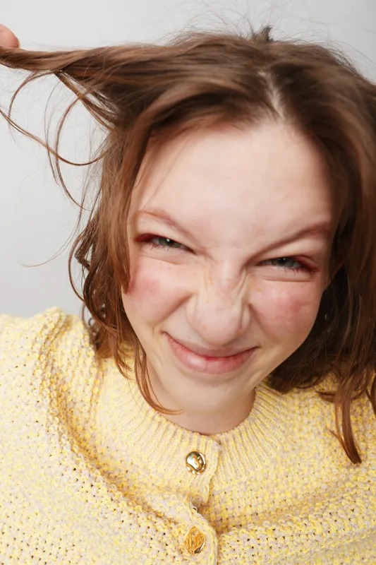 Close-up of a young woman with messy hair pulling strands, making a playful grimace. She wears a yellow knitted sweater against a plain light background. Concept of fun and spontaneity.