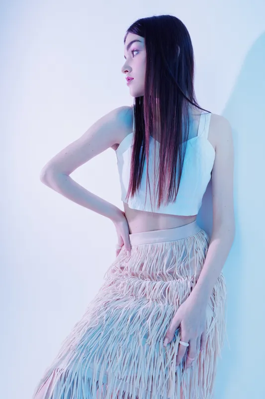 Young woman beauty fashion model poses in a white crop top and fringed skirt against a soft blue background. Concept of style, elegance, and modern clothing trends.