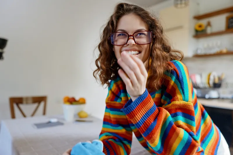 Happy woman in a colorful striped sweater smiles in a cozy home kitchen, enjoying a candid moment, warm light and casual domestic lifestyle in a bright breakfast nook with greenery.