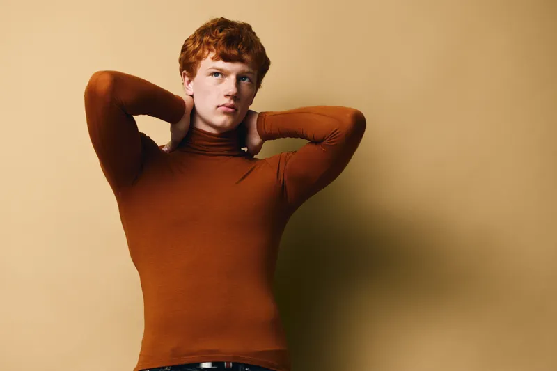 Young man with red hair wearing brown turtleneck sweater posing with hands behind neck against beige background in studio portrait. Stylish casual fashion look for male model.