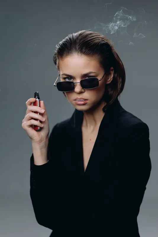 Stylish woman with sunglasses smoking cigarette dressed in black, showcasing attitude and confidence with smoky gray background.