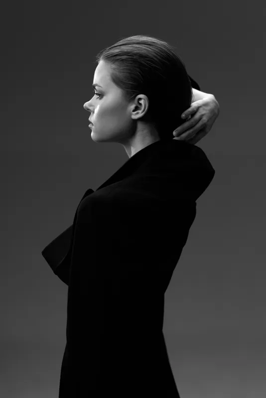 Black and white profile of a young woman in dark clothing with a thoughtful expression and hand raised to head, minimalistic background.