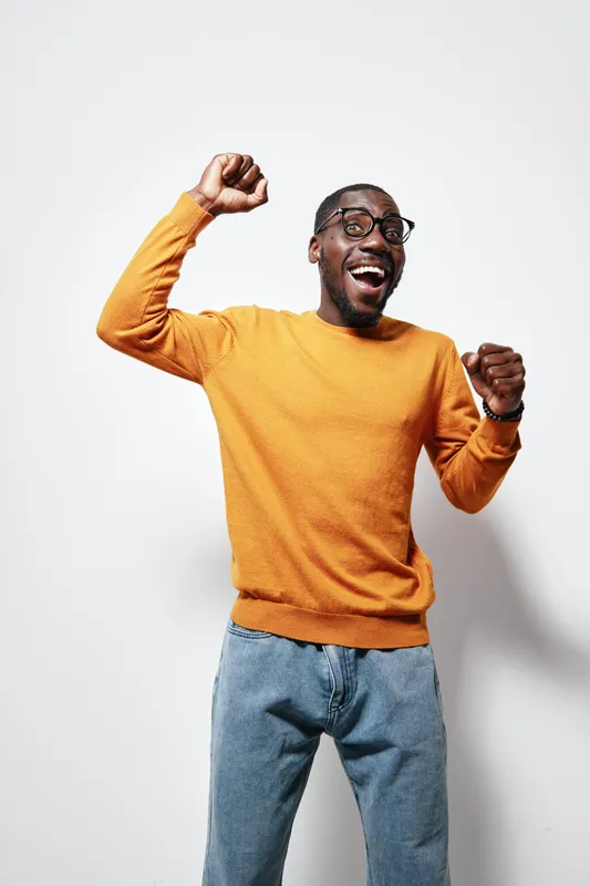 African American man with dark skin wearing glasses and orange sweater celebrating with raised fists and joyful expression against white background in casual outfit.