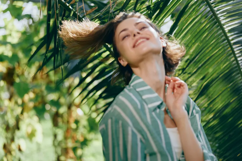 Young woman stands among tropical palm leaves, smiling softly, sunlight framing her face, wearing a striped shirt.