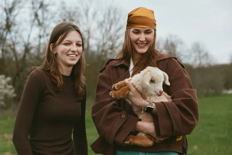 Goat, kid, lifestyle, outdoors, women, friendship, nature, animals reveal two women with a curious goat kid in a sunlit meadow, casual outfits, relaxed posture, candid joyful moment, fresh breeze,
