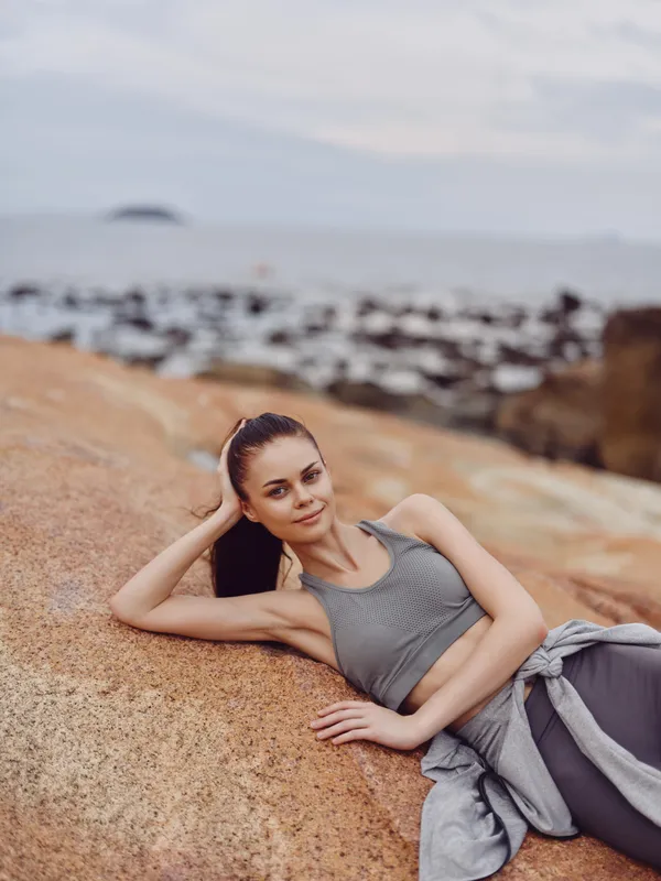 Young athletic woman in gray sportswear resting on rocks by the sea, showcasing a healthy lifestyle and outdoor fitness concept