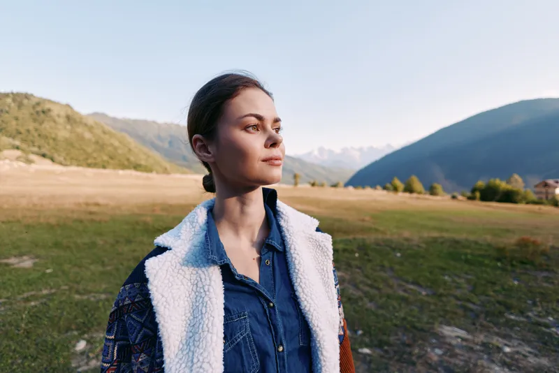 Woman portrait outdoors in mountains and field, nature scene with thoughtful expression and travel mood. Casual denim jacket and coat, serene landscape and soft golden sunlight.