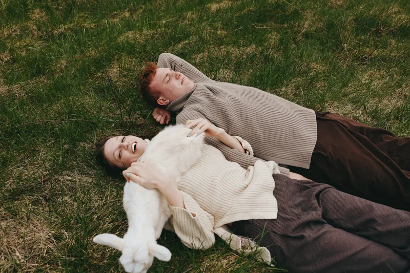 Two friends lie on the grass in cozy sweaters, smiling softly as a fluffy white dog rests nearby. Casual outdoor moment highlighting friendship, comfort, and relaxed wellbeing.