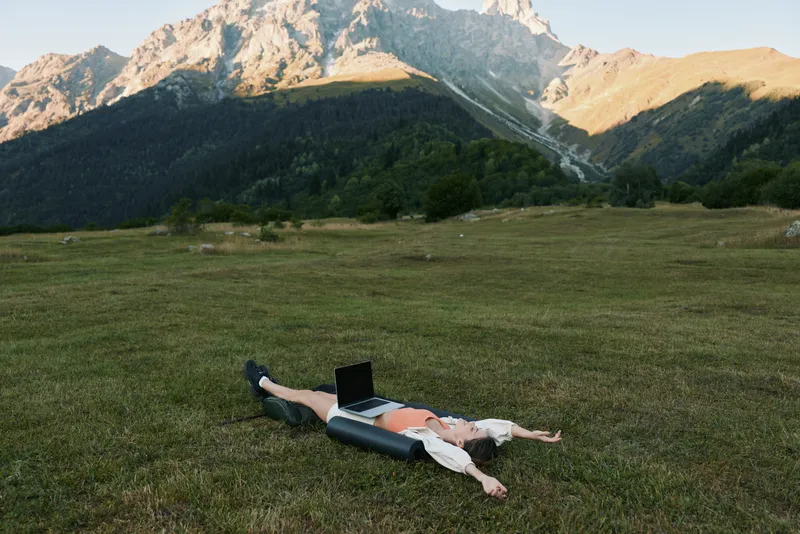A person lies on a grassy meadow with a laptop in a dramatic mountain setting. They appear relaxed yet focused, blending work with outdoor freedom and serene scenery.