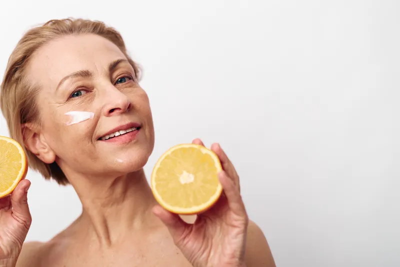 Mature woman with short hair holding two orange halves near face with white cream on cheek. Bright background emphasizes skincare, healthy lifestyle, natural beauty, and vitamin C concept.
