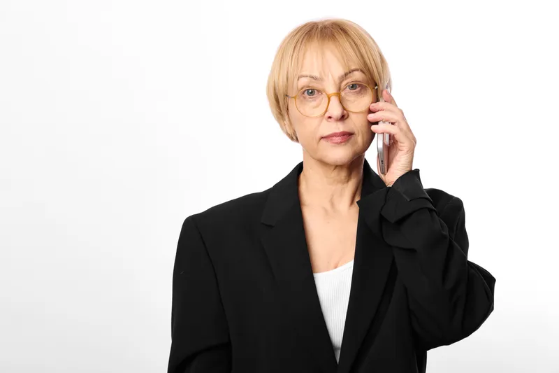 Woman mature portrait with glasses and black blazer, office communication call pose, holding phone to ear, calm confident expression, white background, studio headshot