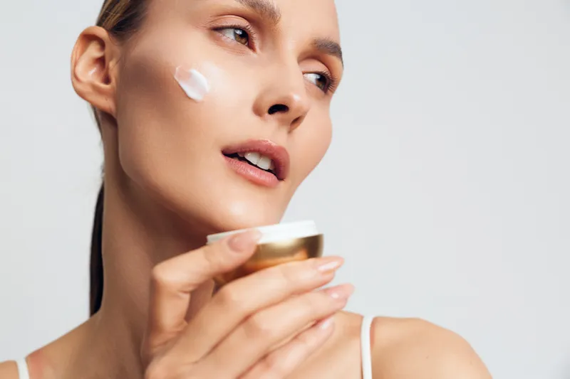 A young woman applies white cream on her cheek while holding a small jar. She looks away with a soft expression against a plain light background. Concept for skincare and beauty routine.