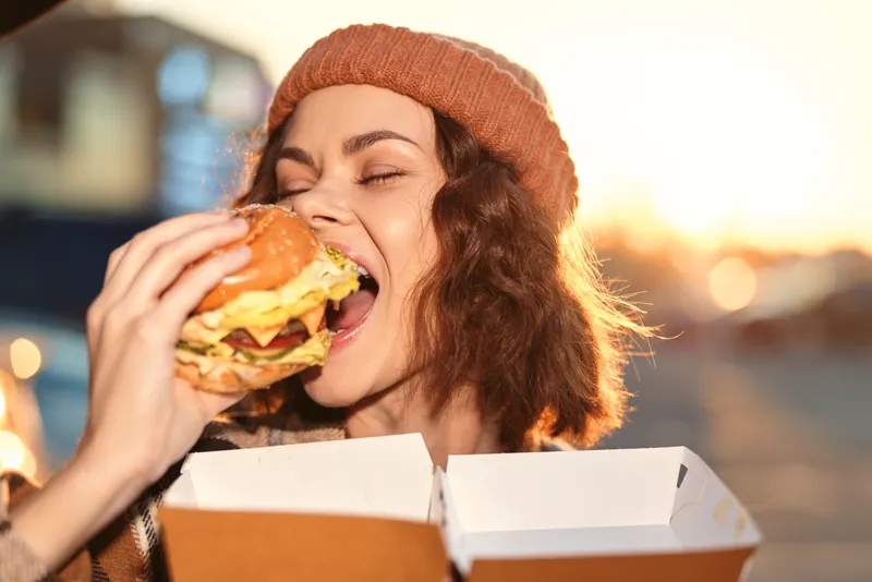 woman burger bite smile outdoor beanie box golden portrait of a joyful woman biting a large burger at sunset, candid lifestyle, authenticity, golden hour glow, emotional storytelling, mindful living