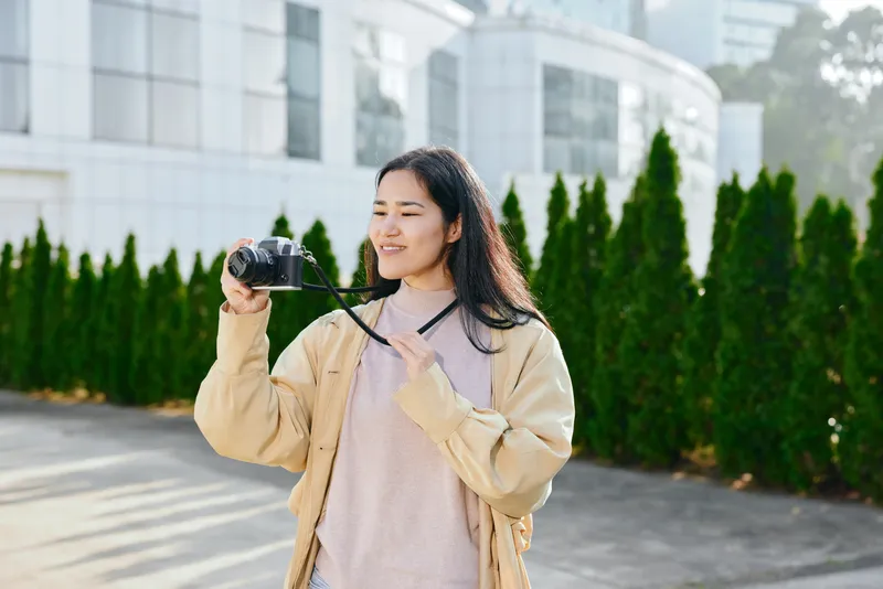 Smiling woman holding a camera outdoors, casual jacket in daylight, urban backdrop with greenery, candid photography moment, lifestyle and travel vibe for stock use