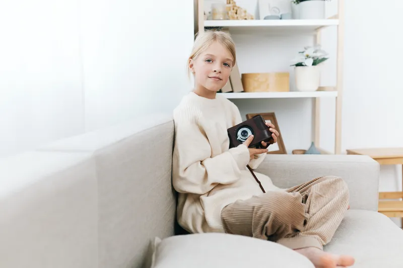 Young girl sits on a light sofa indoors, holding a small camera with a curious smile. Soft natural light and warm tones create a cozy, candid portrait that emphasizes creativity and quiet curiosity.
