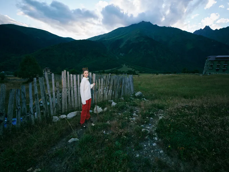 Child standing near a rustic wooden fence in a wide meadow, mountains looming in the background, moody clouds above, peaceful countryside scene with open skies.