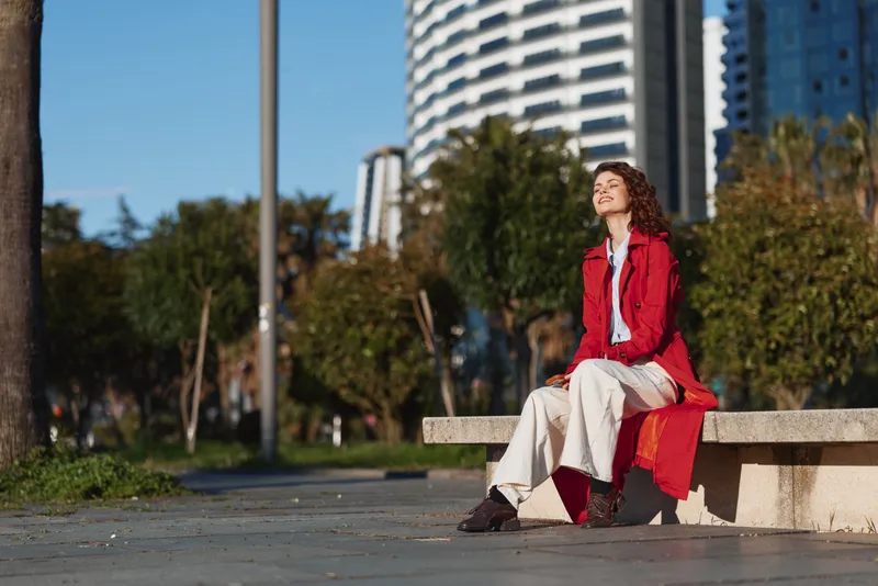 Young woman with curly hair wearing red coat sits on urban bench enjoys sunny day with eyes closed and relaxed smile in modern city park environment.