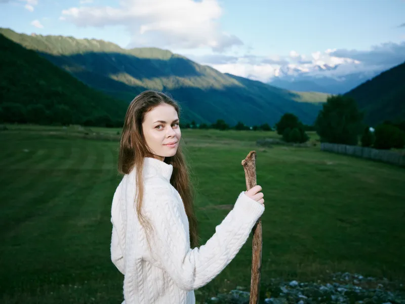 Woman in a white knit sweater holds a wooden staff in a green field with distant mountains, conveying outdoor exploration and serene nature in a peaceful countryside setting