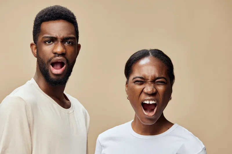 African couple exhibiting strong emotions, expressing anger and frustration, dressed in casual white clothing against a warm beige background