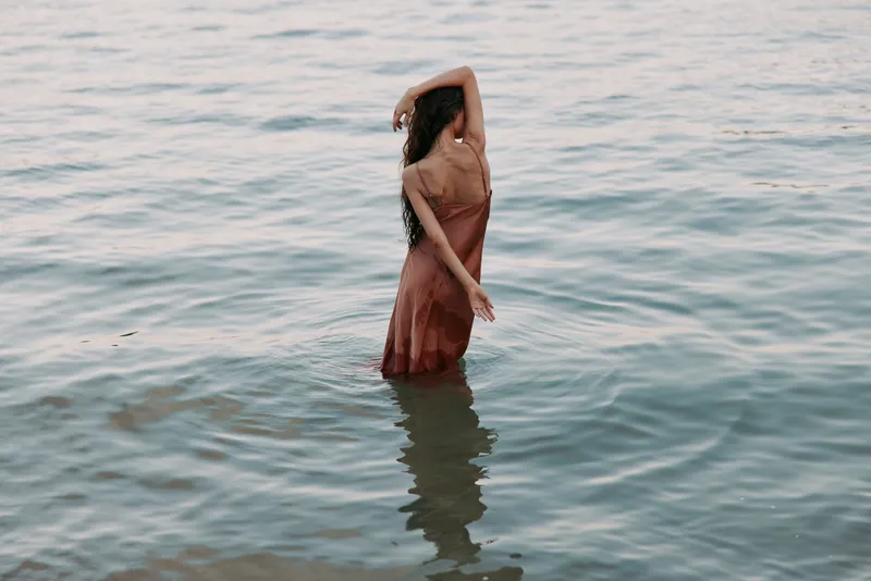 A lone woman stands waist deep in calm water, wearing a flowing dress. Soft light shimmers on the surface, creating a serene, intimate mood.
