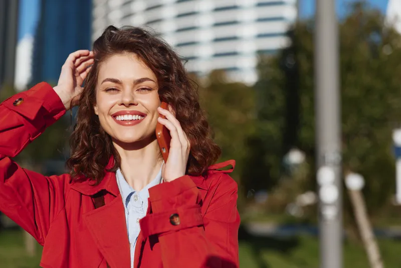 Smiling woman with curly hair wearing a red jacket talks on a smartphone outdoors in an urban park under bright daylight with modern buildings in the background.