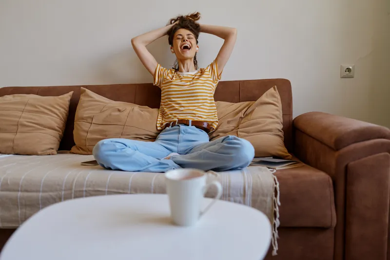Casual woman sitting on a sofa at home, stretching arms with a joyful smile, striped shirt and jeans, mug on a coffee table, warm natural light.
