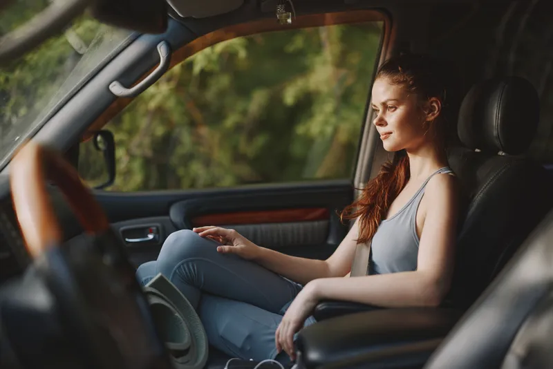 Thoughtful young woman in casual clothing sitting in a car interior, enjoying nature views, reflecting on life with a serene expression