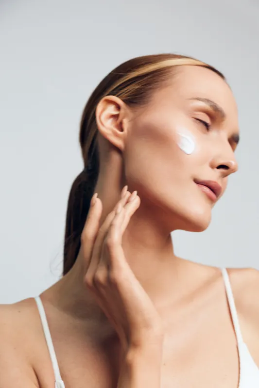 Young woman with clear skin applying white cream on cheek, closed eyes, relaxed expression, natural beauty, soft lighting, minimal makeup, smooth complexion closeup