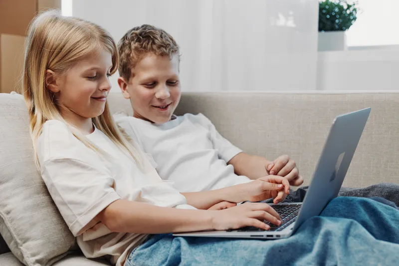 Two children sitting on a couch using a laptop together, smiling and focused. The cozy indoor setting features casual clothing and natural light from a nearby window.