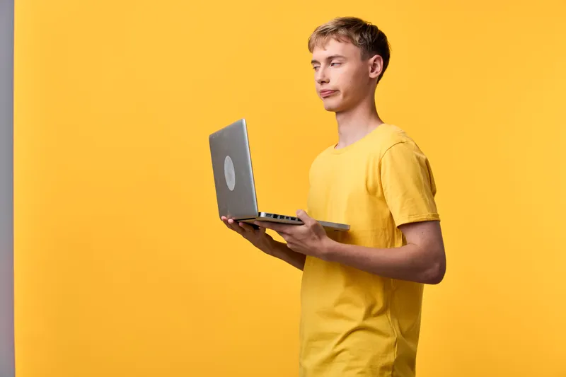 Man in a yellow t shirt using a laptop in a bold studio setting, showcasing digital work, learning, and mobile productivity with focus and modern technology