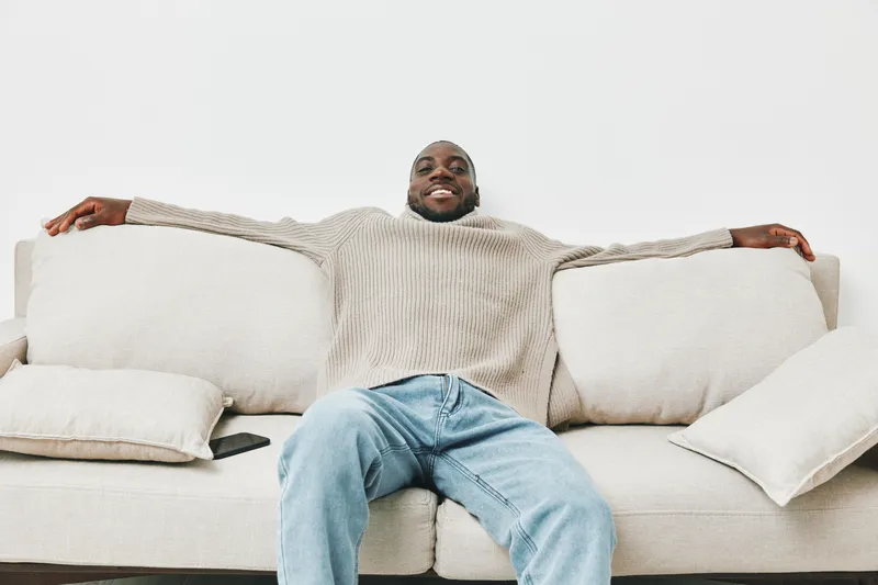 Relaxed Black man in a cozy sweater enjoying leisure time on a sofa, showcasing a sense of comfort and happiness