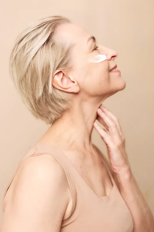 Mature woman with short blonde hair applies face cream on cheek, smiling gently. Skin care, anti aging and self care concept with soft natural light and beige studio background.