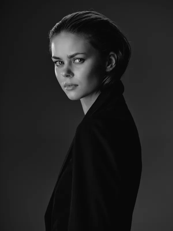 Black and white portrait of a serious young woman with slicked-back hair wearing a dark jacket on a dark background.