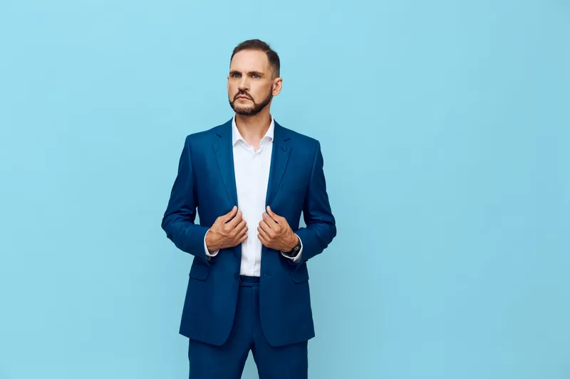 A professional man in a tailored suit stands against a solid blue backdrop, exuding confidence and focus as he adjusts his blazer for a business moment.