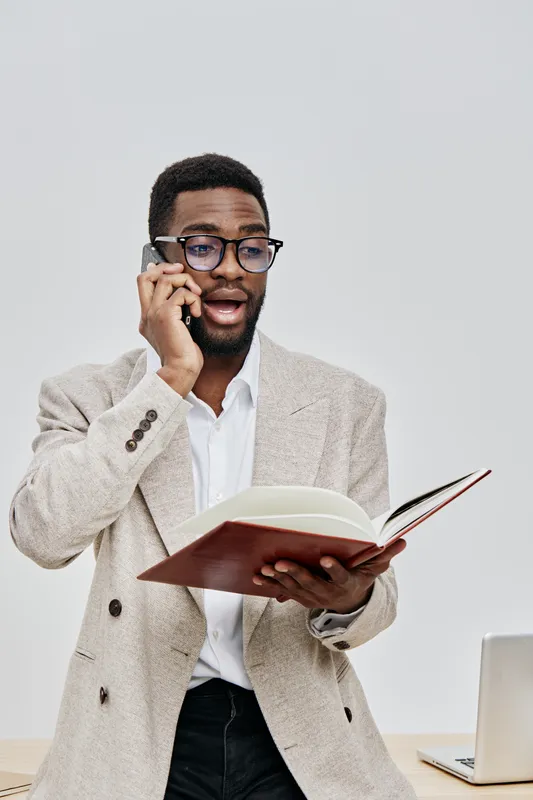 Professional young man of African descent wearing glasses and beige blazer talking on phone while holding open notebook, standing in office environment with neutral background. Business communication 