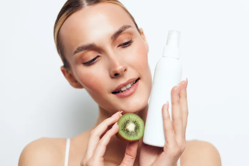 A young woman with clear skin holds a white cosmetic bottle and a halved kiwi fruit, smiling gently. The bright studio background highlights natural beauty and skincare concepts.