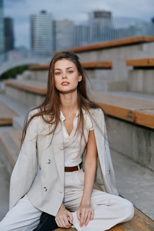 Young woman in beige business suit seated outdoors with urban cityscape background, confident expression, natural makeup, and stylish casual look