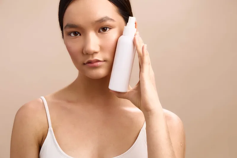 Asian woman holds a cosmetic bottle near her temple in a calm bathroom, showcasing a skincare routine. She looks focused and confident, conveying a serene selfcare mood with modern elegance.