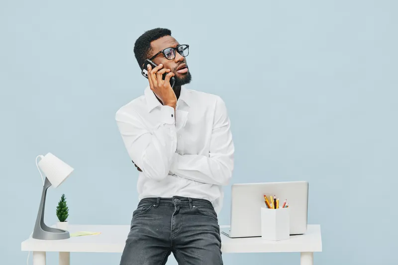 Confident young African American man wearing white shirt and glasses talking on mobile phone in modern office with laptop, desk lamp, and minimal decor, studio shot, professional lifestyle concept